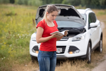 Portrait fo young woman got stuck on countryside road because of her broken car reading manual book