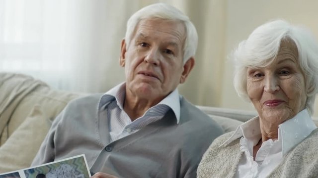 Tilt up of happy senior couple with grey hair looking at family photo album and chatting, then posing for camera