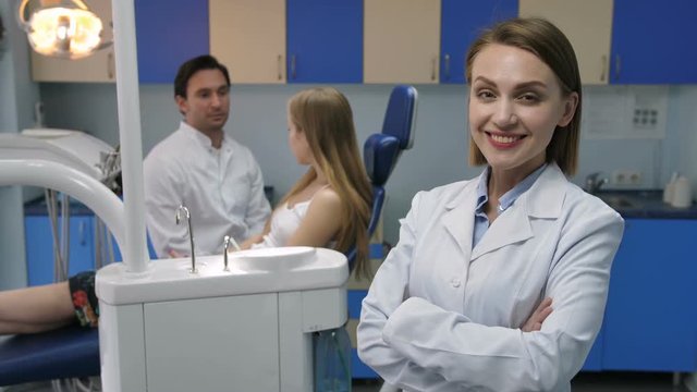 Pretty mid adult female dentist standing and smiling to camera with arms crossed in lab coat. Male dental doctor's assistant talking to female patient in dental chair at background at dental office