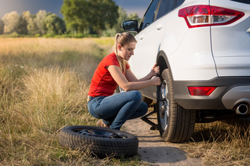 Naklejka na ściany i meble Young woman changing car spare wheel on the deserted road at field