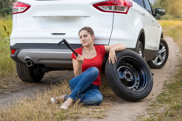 Young woman sitting at broken car in field and holding car jack