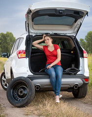 Upset young woman sitting in open trunk of broken car in field