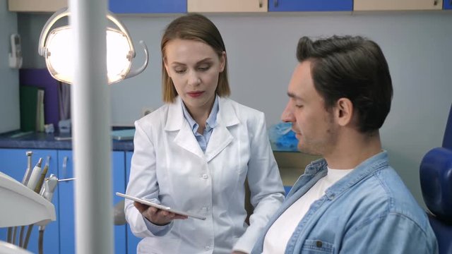 Beautiful female dental doctor in white laboratory coat showing young male patient his dental radiography x-ray shot. Woman dentist discussing diognosis and treatment with young adult man client