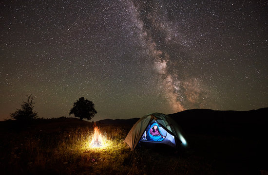 Woman hiker having a rest at night camping beside campfire in the mountains under incredible beautiful starry sky and Milky way. Girl sitting inside illuminated tent and sleeping bag. Astrophotography