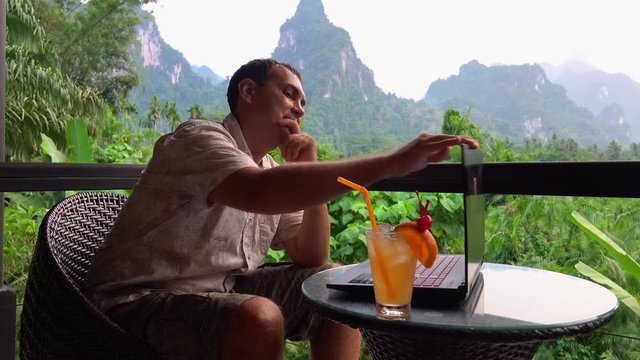 Male Traveler Using Laptop with Scenic Limestone Rocks View. Digital Nomad Working on Terace. Freelancer Typing on Computer in Asia. Remote Work in Thailand