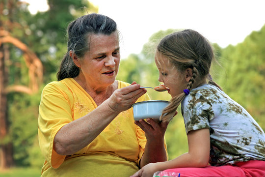 A grandmother is feeding her granddaughter by spoon from bowl outdoors