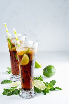 Refreshing alcohol beverage cuba libre with cola, ice, mint, lime, straw in two wet long glasses in modern white kitchen interior, copy space.