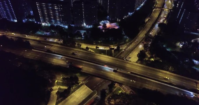 Cars driving on freeway interchange at night. Tilting drone view of highway lit up at night next to city scape. Dramatic batman lighting, action movie vibe.