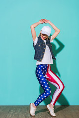 stylish young woman in cap, white shirt, denim jacket and leggings with american flag pattern posing on blue background