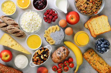 Healthy breakfast ingredients, food frame. Granola, egg, nuts, fruits, berries, toast, milk, yogurt, orange juice, cheese, banana, apple on gray background, top view, copy space.