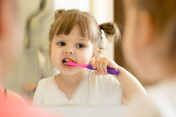 Cute child girl looking at mirror using toothbrush cleaning teeth in bathroom every morning and...