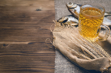 Foam beer in a mug, dried fish stockfish and rye ears wrapped in burlap cloth on a burnt wooden table background with copy space.