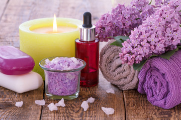 Fototapeta na wymiar Soap, burning candle, bowl with sea salt, red bottle with aromatic oil, lilac flowers and towels on wooden boards