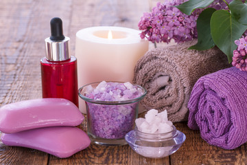 Obraz na płótnie Canvas Soap, red bottle with aromatic oil, burning candle, bowls with sea salt, lilac flowers and towels on wooden boards