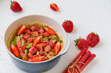 Raw rhubarb strawberry pie ready for cooking in the baking dish on the white kitchen table. Decorated with fresh ingredients for summer vegan pie. Side view