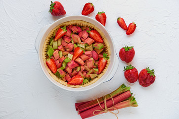 Raw rhubarb strawberry pie ready for cooking in the baking dish on the white kitchen table. Decorated with fresh ingredients for vegan summer pie. Top view