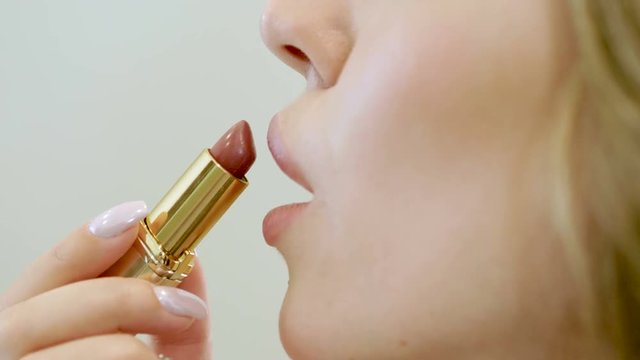 Face make-up. Close-up shot of young beautiful blonde woman applying a beige lipstick on her lips. 4K