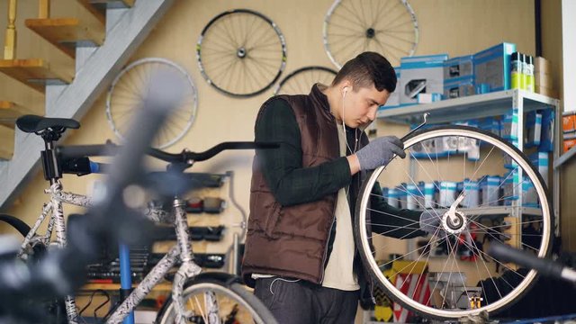 Self-employed mechanic is repairing bicycle wheel with wrenches professional instruments while working in small workshop. Young man is listening to music with earphones.