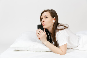 Calm young woman lying in bed with white sheet, pillow, blanket on white background. Beauty female listening music from earphones in mobile phone, spending time in room. Rest, relax, good mood concept