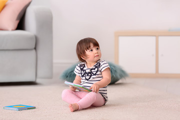 Cute baby girl with book sitting on floor at home