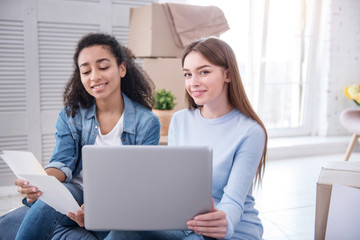 Sharing ideas. Upbeat young girls sitting on the floor and posing for the camera while shopping online for wall paint, having moved in to a new apartment