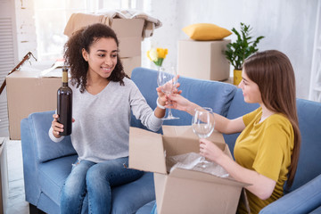 Home-warming party. Happy young girls sitting on the sofa and unpacking two wine glasses for celebrating moving into a new flat