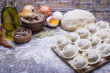 Homemade dumplings of their dough and minced meat, a set of products for cooking: flour, dough, minced meat, eggs, olive oil, spices, salt, pepper, bay leaf on a wooden board, background.