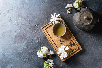 Traditional ceramic cup of hot green tea on wooden board with black iron teapot, spring flowers white magnolia and cherry blooming branches over dark blue texture background. Top view, copy space. © Natasha Breen