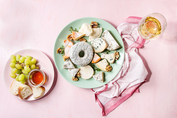 Cheese plate assortment of french cheese served with honey, walnuts, bread and grapes on turquoise ceramic plate and glass of white wine over pink pastel background. Top view, space.