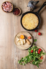 Spanish potato omelette tortilla with bacon served in cast-iron pan with sauce and ingredients above over wooden background. Top view, space.