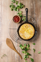 Spanish potato omelette tortilla with bacon served in cast-iron pan with tomato sauce and field salad above over wooden background. Top view, space.