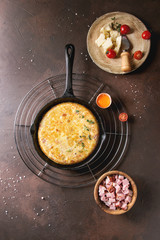 Spanish potato omelette tortilla with bacon served in cast-iron pan on cooling rack with ingredients above over over dark brown texture background. Top view, space.