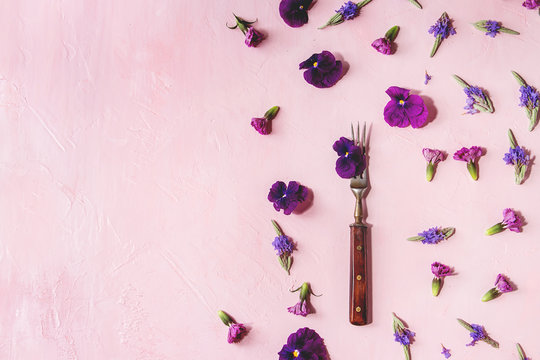 Variety of purple edible flowers for dish decorating with vintage fork over pink pastel background. Top view, space.