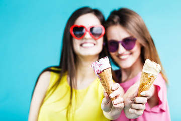 Two pretty happy smiling girls dressed in colorful t-shirts and sunglasses have fun with icecream on blue background, carefree youth concept