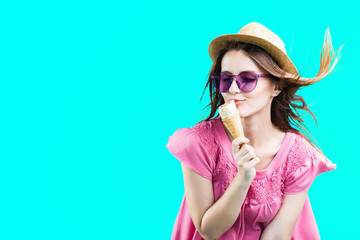 Fototapeta premium Pretty blonde woman dressed in rose shirt, hat and sunglasses eating wafle icecream before blue background