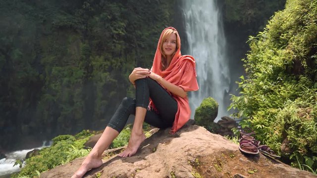 Young caucasian woman in head scarf sitting in front of Bajing Waterfall on sunny day in Pekalongan,Indonesia