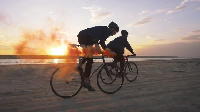 Two young men riding bicycles on the beach with orange smoke during sunset