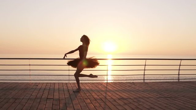 Beautiful silhouette of ballerina in ballet tutu and point on embankment above ocean or sea at sunrise. Young beautiful long haired woman practicing and exercises. Morning bosk