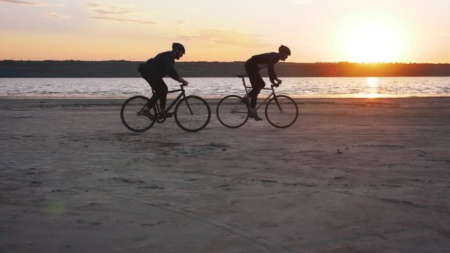Two young men riding bicycles on the beach on the background of an orange sunsetting sky