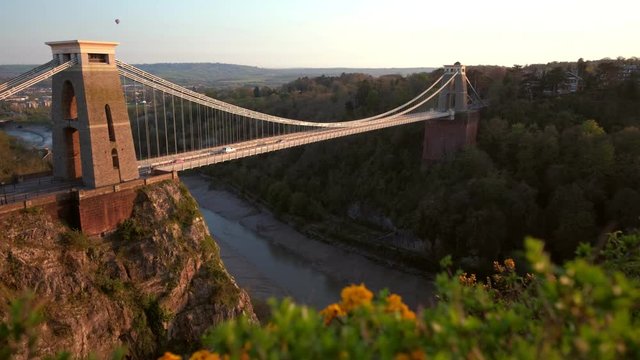 Camera slides over hedgerow with the Clifton Suspension Bridge behind. Shot during a spring sunset with a hot air ballon in the sky