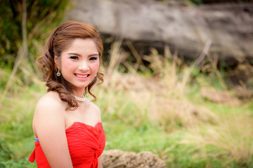 Close up Beautiful woman wear red evening dress happy smile sitting on a rock in the woods.