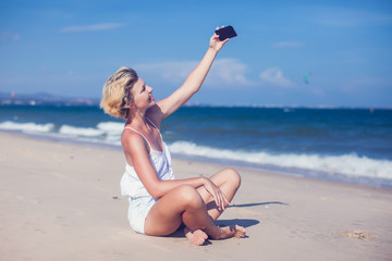 Smiling young woman take a selfie photo at sandy beach by the sea at sunset