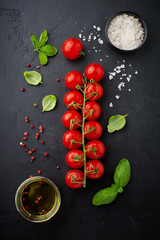 Branch of raw fresh cherry tomatoes, olive oil and leaves basil on black background. Food background. Selective focus. Top view.