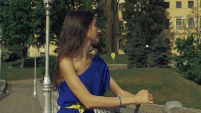 Stunning long brown hair woman in trendy dress leaning on the bannister of pedestrian bridge and looking away at cityscape with enigmatic smile, remembering great moments while relaxing outdoors.