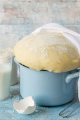 Homemade dough in a small blue saucepan, next to a glass with milk and flour on an old wooden table. 