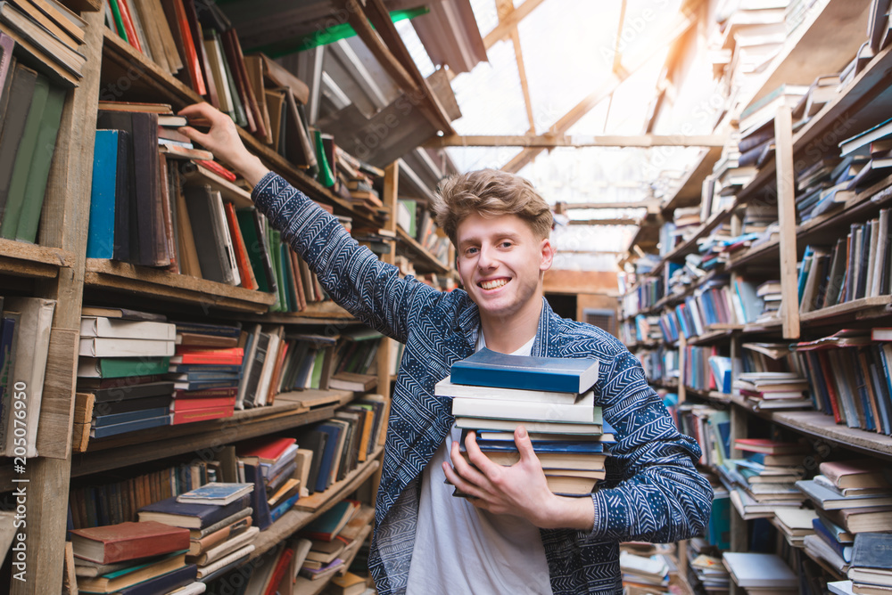 Wall mural Happy student with a lot of books in his hands is in the library, takes books from the shelf and looks at the camera with a smile. Portrait of a smiling young man in an old bible. Learning the concept