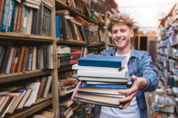 Portrait of a young student offering a book. A man with lots of books in his hands is in the library and smiles. Concept of training. Focus on books