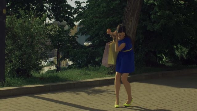 Elegant beautiful woman with shopping bags in blue mini dress and yellow high heels twisting her ankle while walking on city street. Gorgeous woman touching foot in pain due to sprained ankle.