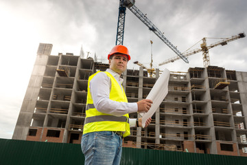 Portrait of male engineer in hardhat and safety with blueprints standing against working building cranes
