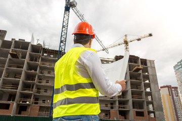 Rear view image of male architect in hardhat and vest looking at working building cranes